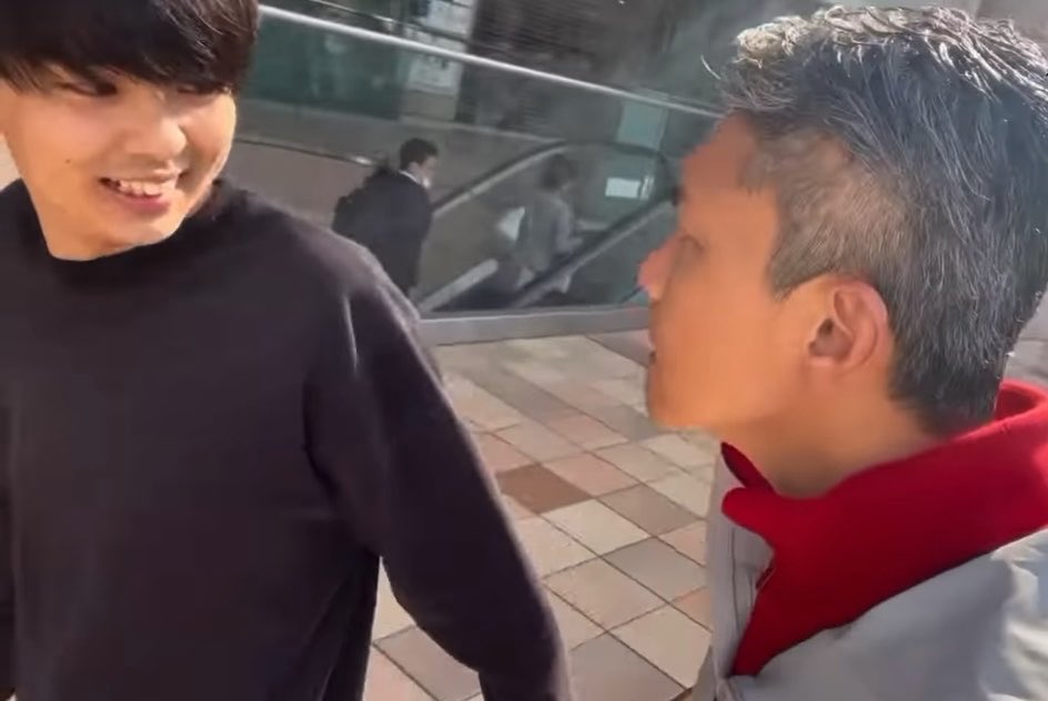A bald student in a red and gray jacket confronts a smiling dark-haired student in a black sweater on a tiled walkway near an escalator, illustrating the tense moment before the famous delinquent picked a fight with the wrong person.