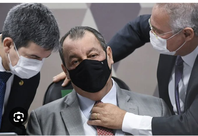 Brazilian politician Omar Aziz wearing a black mask adjusts his tie while seated between two men also wearing masks, likely during a Senate hearing.