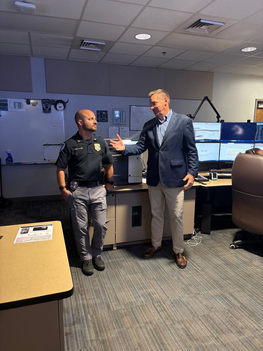 Two men stand in a police operations room. One wears a police uniform, while the other wears a blazer and gestures as they speak. Desks, computers, monitors, and wall displays surround them. Some text is visible on the displays and papers.