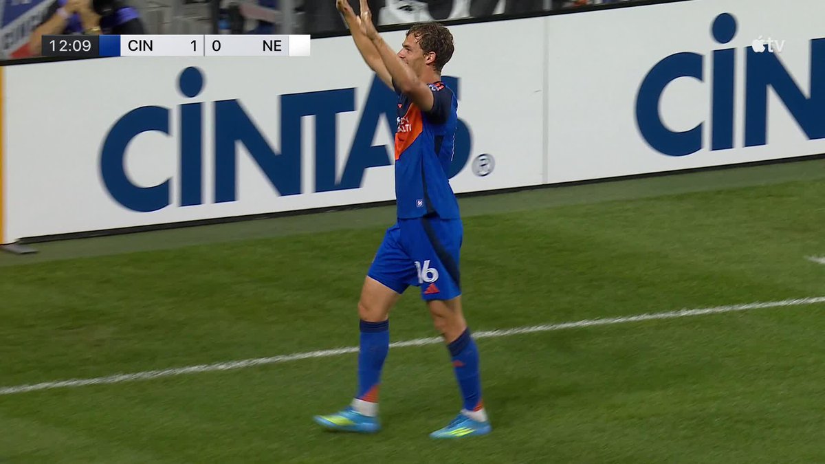 A player wearing a blue Cincinnati jersey with the number 16 raises his hands in celebration on the field after scoring a goal. The scoreboard shows "CIN 1 - 0 NE" at 12:09, confirming FC Cincinnati's early lead as mentioned in the post.
