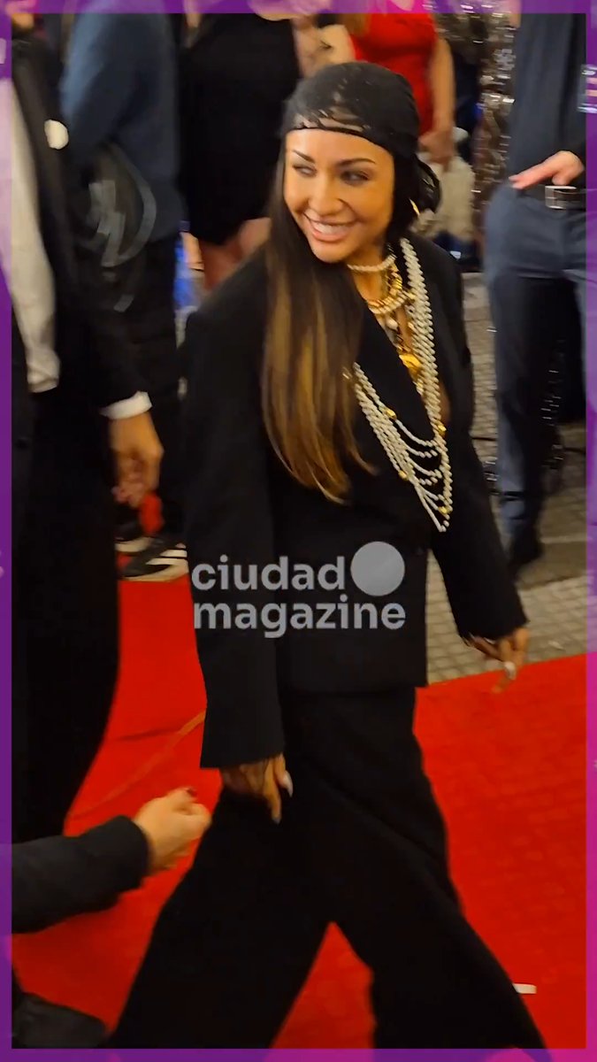 A celebrity photo with a 'ciudad magazine' watermark showing a smiling woman in black attire, a patterned headscarf, and layered pearl necklaces walking on a red carpet.