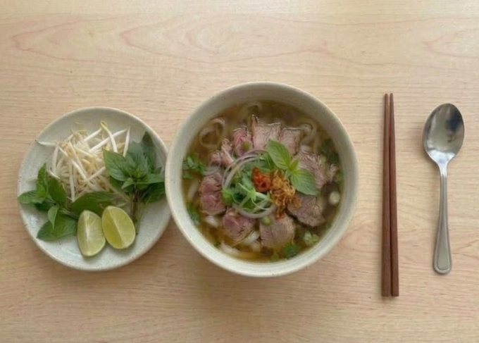 A bowl of Vietnamese pho with sliced beef, onions, and herbs sits on a wooden table alongside a plate of bean sprouts, lime wedges, and Thai basil, with chopsticks and a spoon to the right.