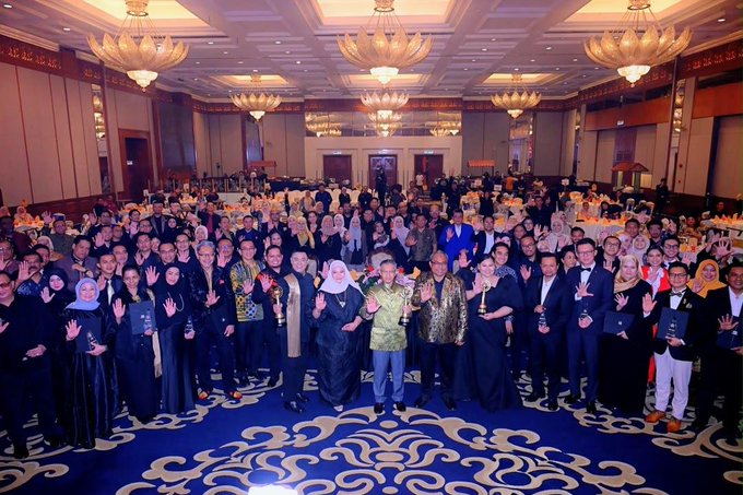 A large group photo of attendees at a formal gala event, with people waving at the camera in an ornate ballroom with chandeliers and a blue patterned carpet.