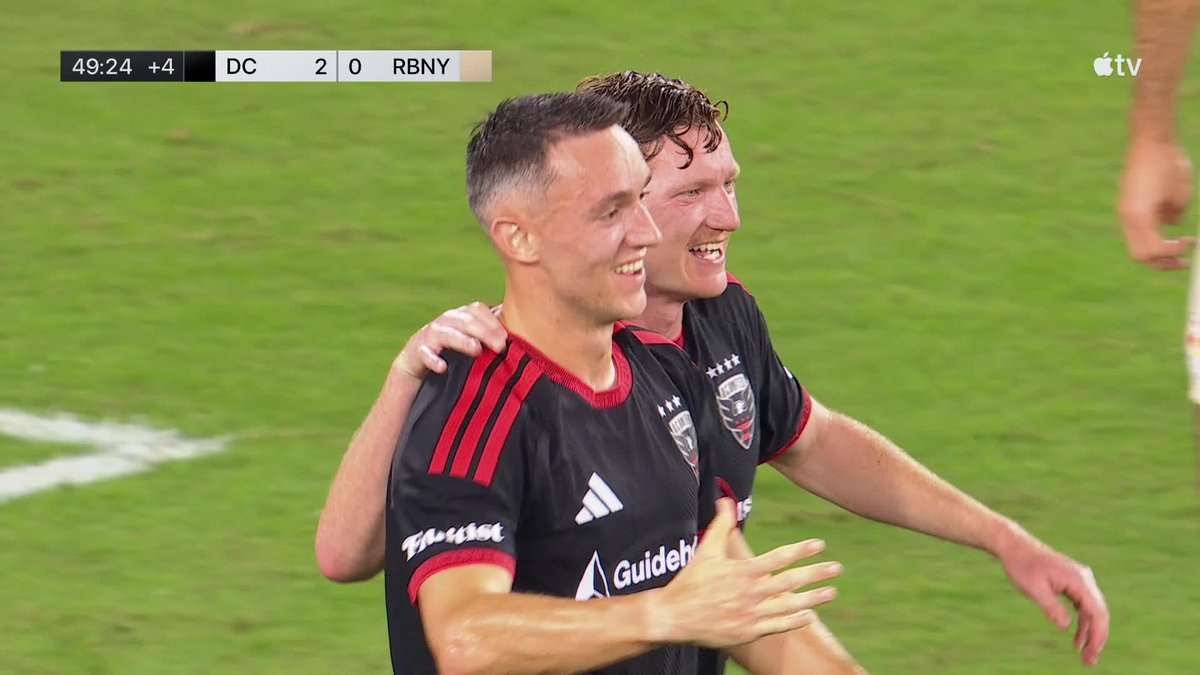 Two soccer players in black D.C. United jerseys with 'Guidehouse' and 'Flojet' sponsor logos celebrate on the field; the scoreboard shows 49:24 +4 with DC leading RBNY 2-0 on an Apple TV broadcast.