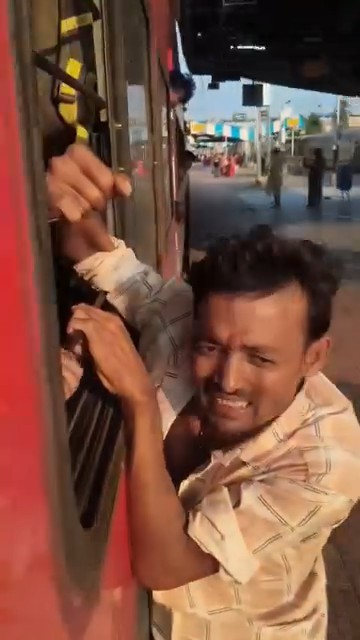 A man in a striped shirt is clinging to the side of a red train, gripping the window frame with a strained expression as the train appears to be moving through a station.