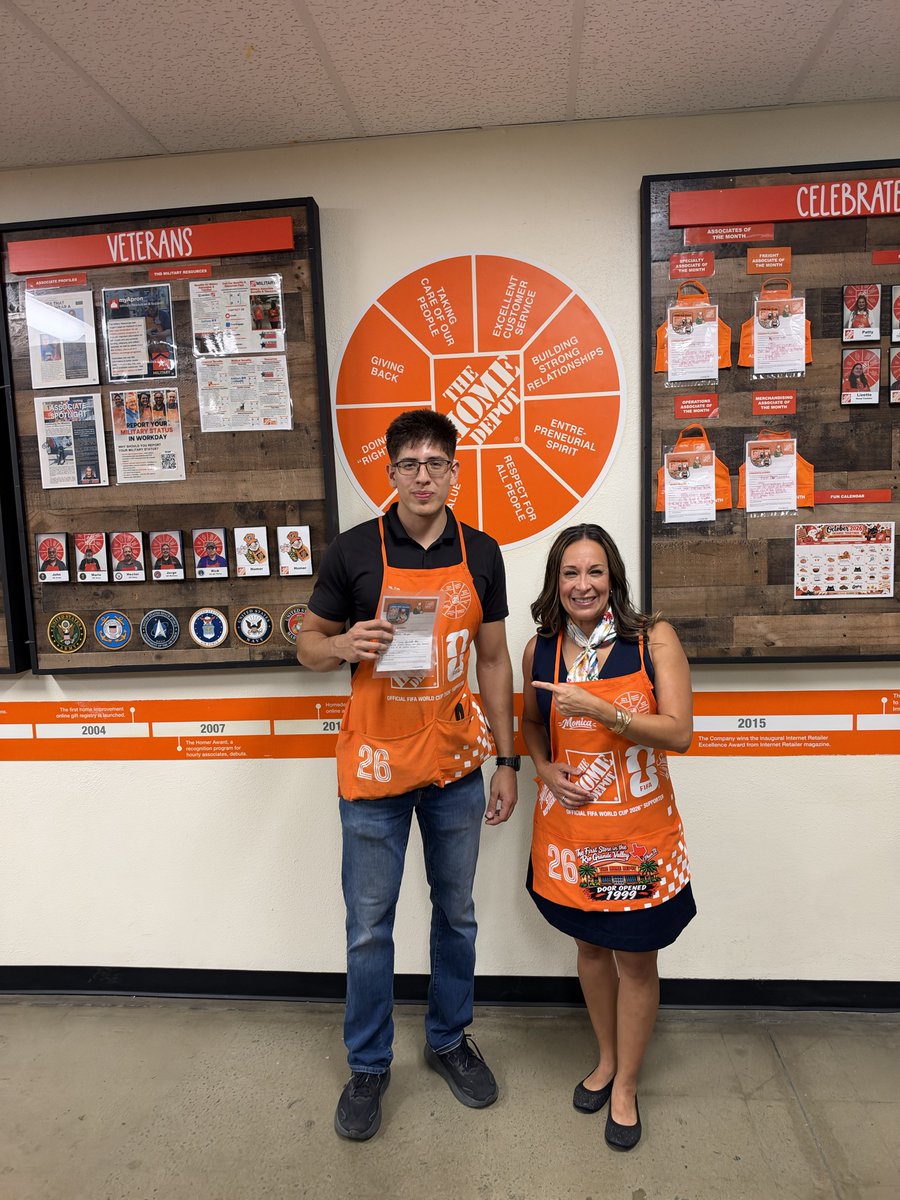 Two people wearing orange aprons pose in front of wall displays and an orange circular values graphic. The man holds a paper, while the woman points toward him. There is text throughout the displays and on the aprons.