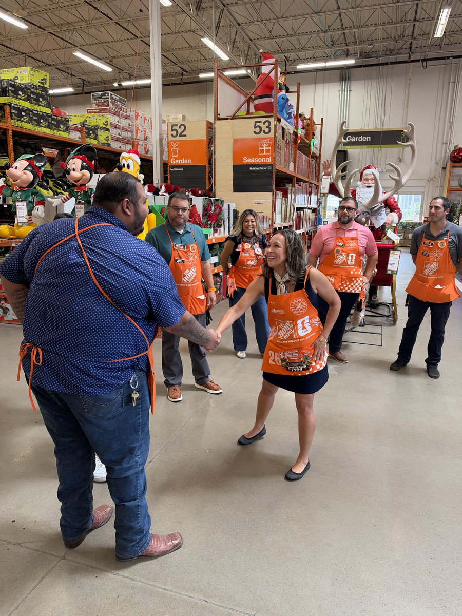 A group of retail employees wearing orange aprons gather in a store seasonal section. A woman and a man shake hands while coworkers look on. Holiday character displays and store shelving are visible behind them, along with signs and other text.