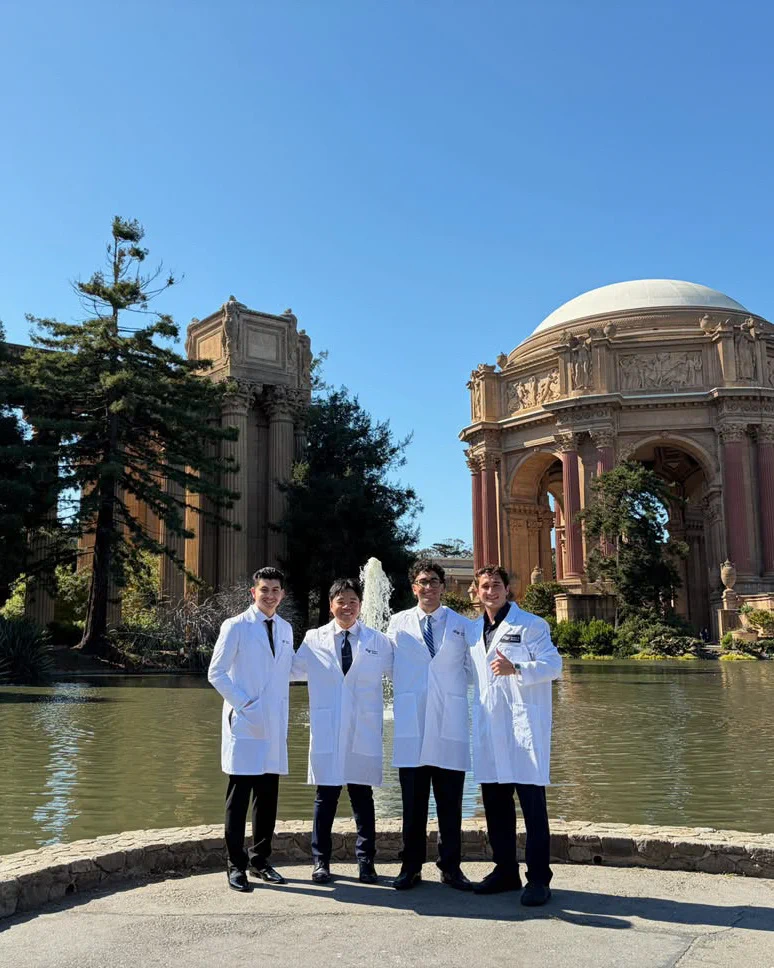 Four men in UCSF School of Dentistry white lab coats stand smiling by a pond with the Palace of Fine Arts in the background on a sunny day.