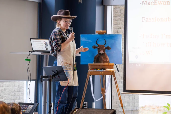 Artist wearing a cowboy hat and apron presents a painting of a bull on an easel.