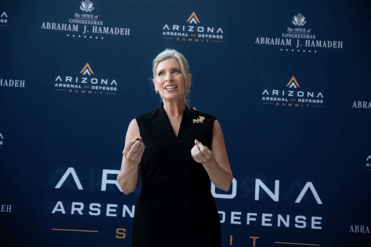 A woman with short gray hair, wearing a sleeveless black dress and a gold brooch, speaks with her hands raised in front of a dark blue event backdrop. The backdrop contains repeated logos and text.