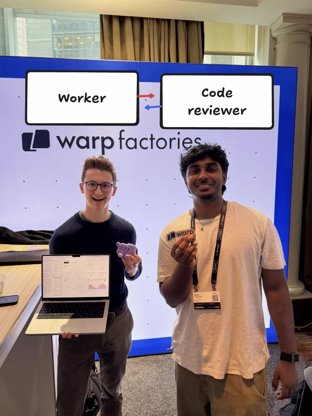 Two people at a Warp Factories booth. One holds a laptop showing an analytics dashboard and a small purple plush toy, and the other holds up a Warp sticker. Above them, labels reading "Worker" and "Code reviewer" have arrows pointing back and forth between them.