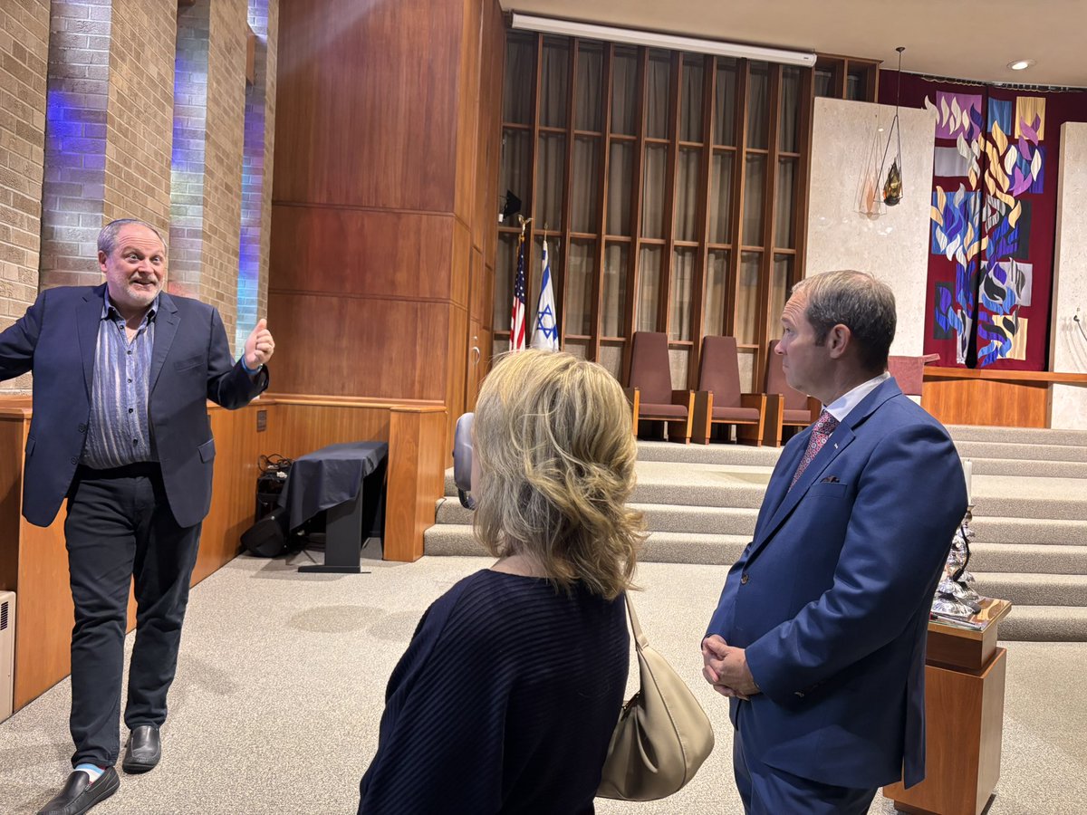 Three adults are gathered inside a synagogue or similar worship space. A man on the left gestures as he speaks, while a woman and a suited man listen. A raised platform, flags, seating, and colorful decorative panels are visible in the background. No prominent text is visible.