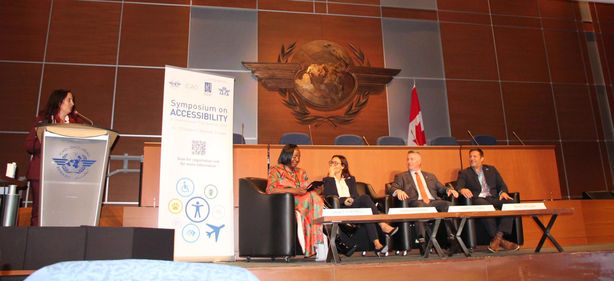 A panel of five people is seated on a stage with the backdrop of a large emblem on a wood-paneled wall. A person stands at a podium on the left, and a Canadian flag is visible on the right. A banner next to the panel reads "Symposium on Accessibility" with various logos and icons, including the ICAO and CAD logos.