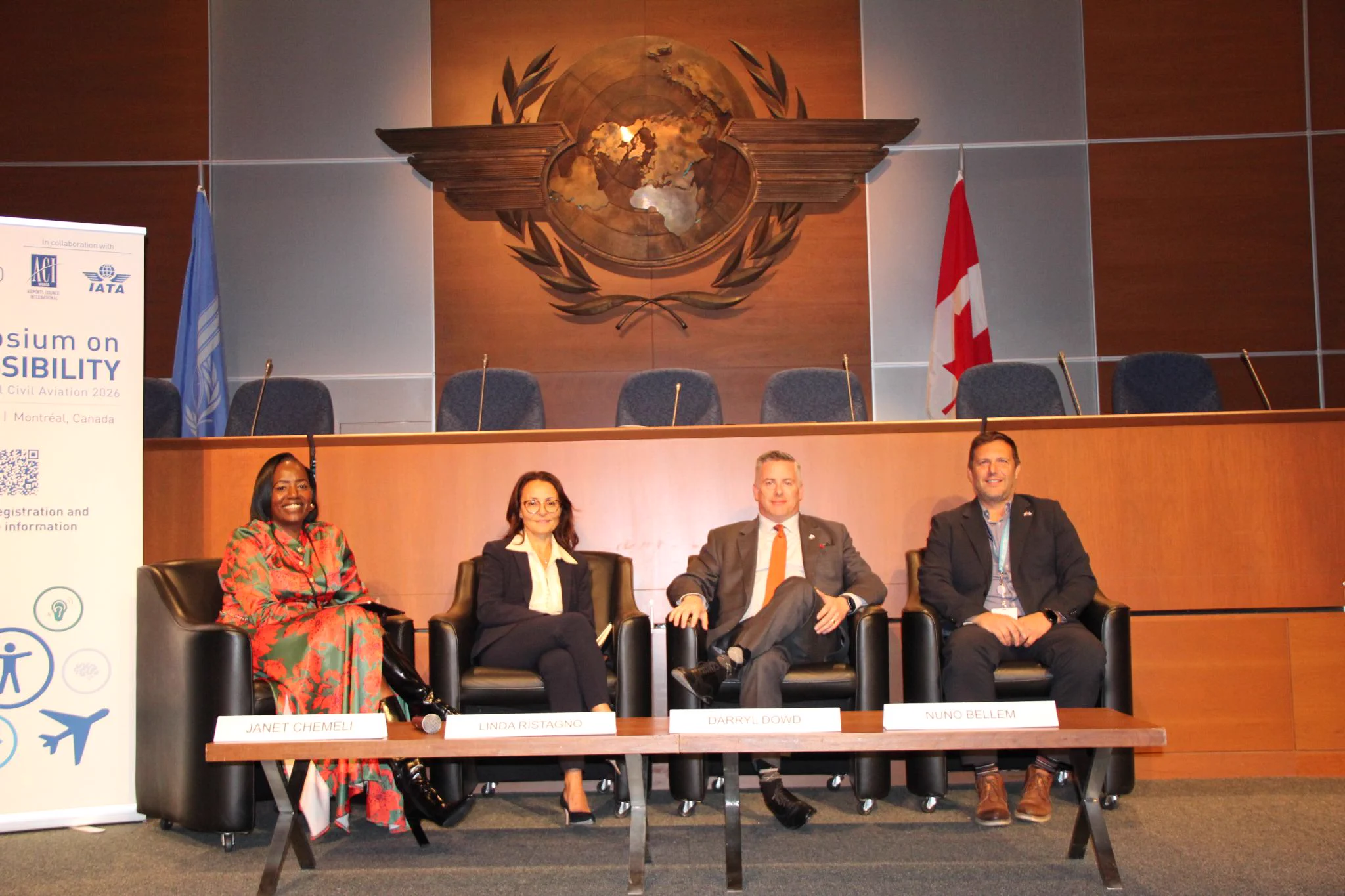 Four individuals are seated on a panel in a formal setting. Behind them is an emblem resembling a world map with wings. A banner on the left includes text and logos related to a symposium on sustainability in Montreal, Canada. A Canadian flag is visible in the background.