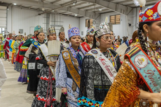 Line of Tribal princess with crowns, sashes, in like walking to grand entry in a expo center