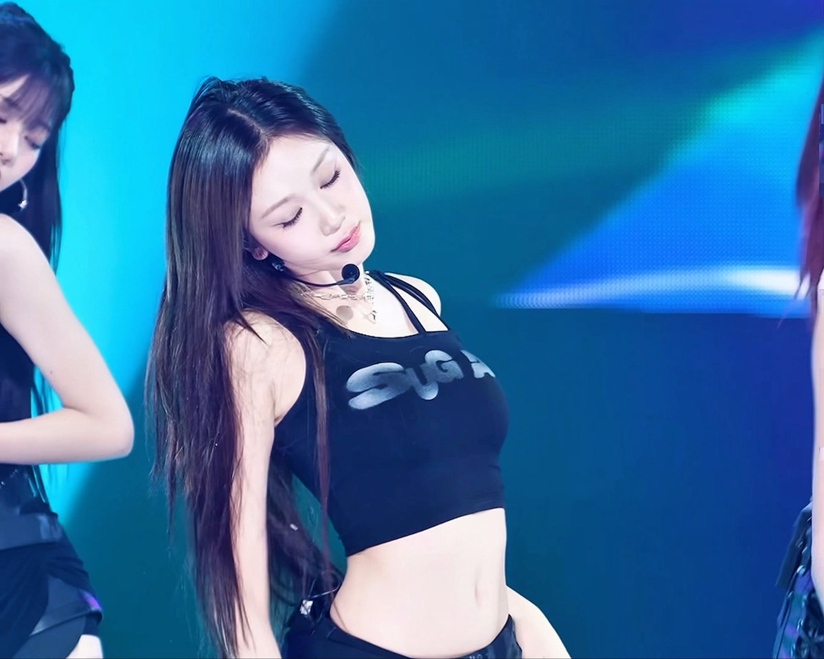 A female K-pop performer with long dark hair dances onstage under blue lighting, wearing a black crop top, headset microphone, and layered necklaces.