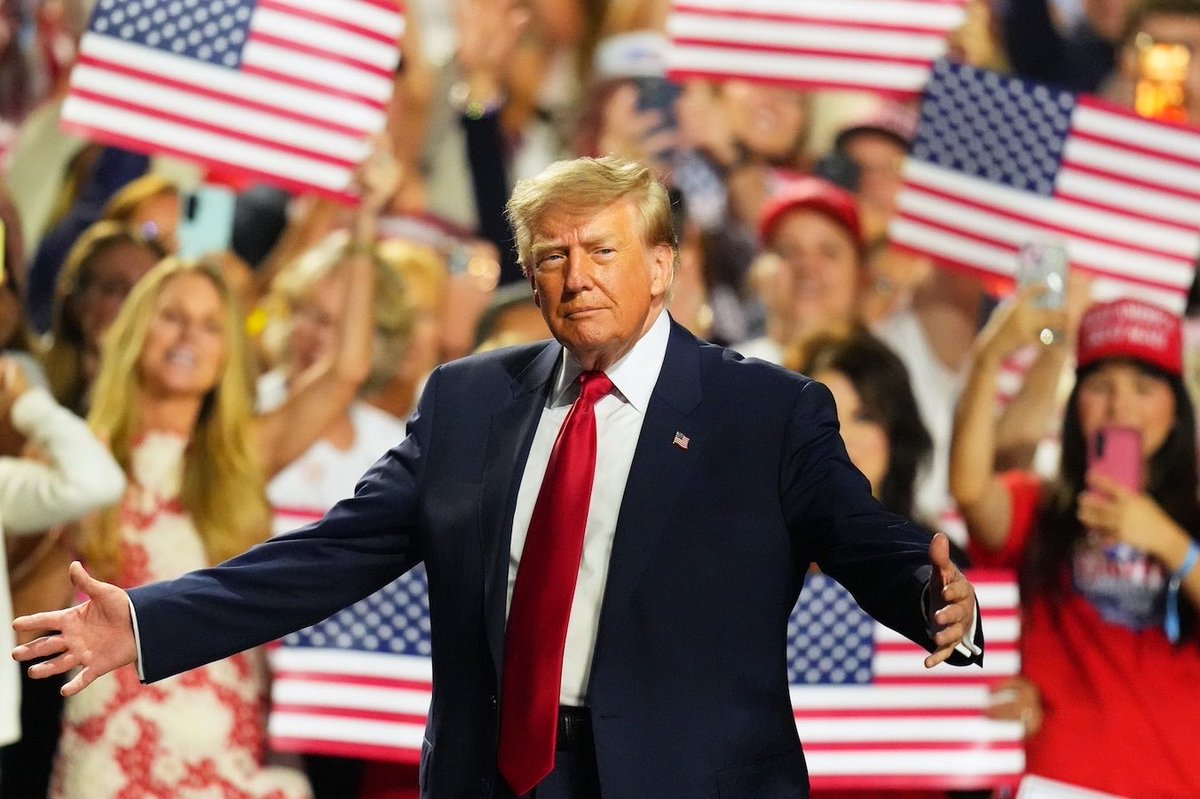 A man in a dark suit and red tie stands with his arms outstretched in front of a cheering crowd holding American flags. There is no prominent text.