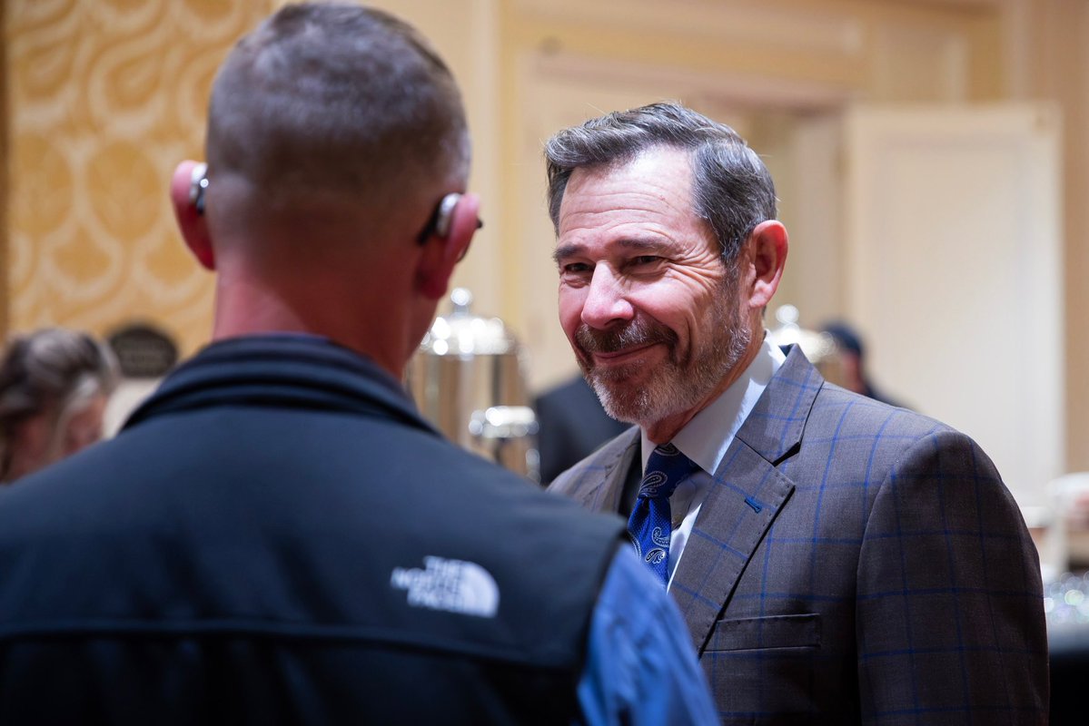 A man in a gray checkered suit smiles while speaking with another man seen from behind at an indoor event. Other people and serving equipment are visible in the background. There is a small logo on the back of the foreground man's vest.