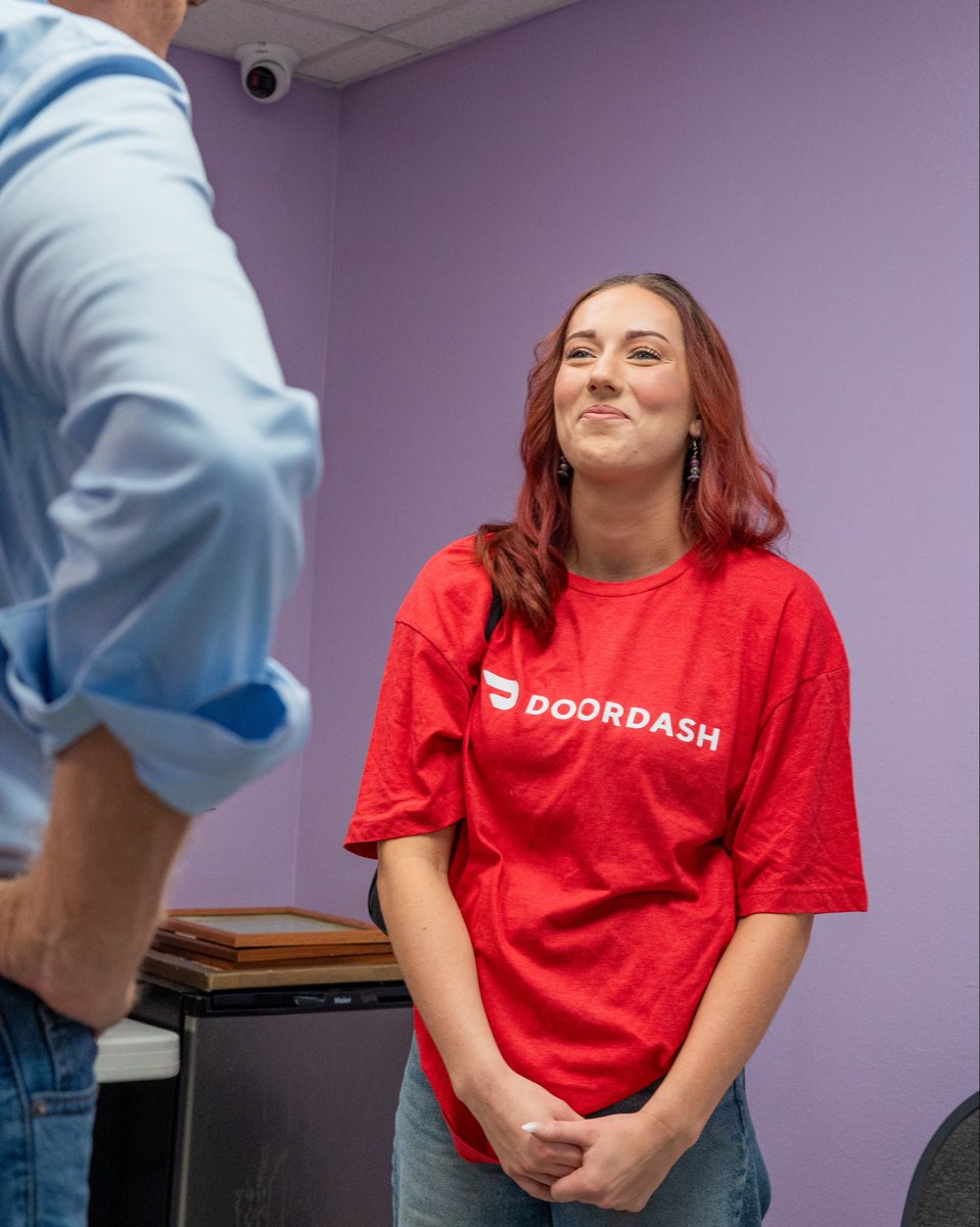A red-haired woman wearing a red T-shirt stands in a purple-walled room, speaking with a partially visible person in a light blue shirt. A security camera is mounted near the ceiling. Text appears on the woman's shirt.