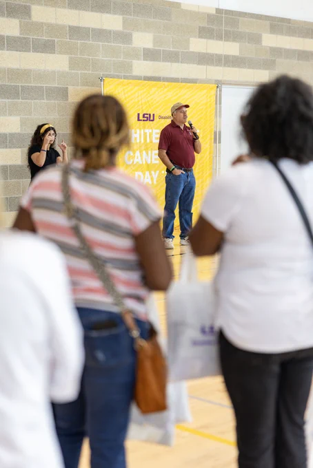 A person is speaking into a microphone in front of a yellow LSU banner that reads "White Cane Day." A sign language interpreter stands nearby. Several attendees are in the foreground, facing away.