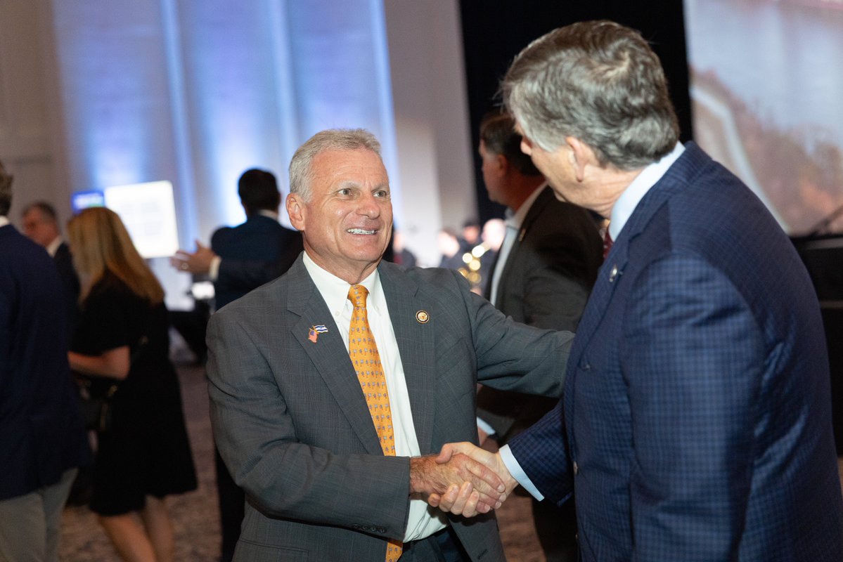 A photograph of two men in suits shaking hands and talking at an indoor event. Other attendees and a large display are visible in the background, with some text present.