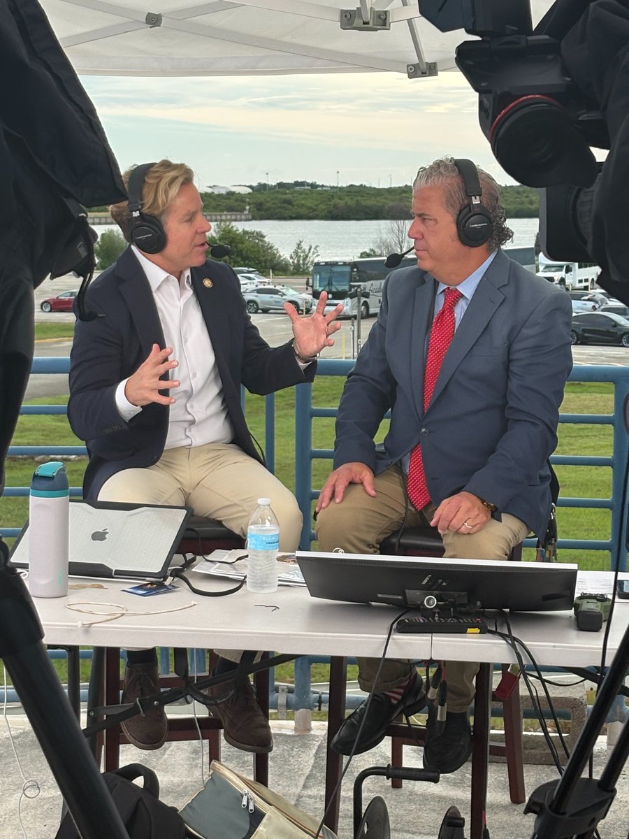 Two men wearing headsets sit at a broadcast setup and talk while cameras and equipment record them. They are seated outdoors near a body of water and a parking area. There is some text on equipment and papers.