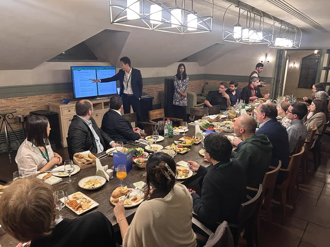 David Dao points to a document on a screen as guests listen around the dinner table, with blue PACE booklets among the shared dishes. Veena Balakrishnan stands nearby.