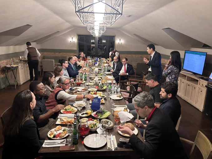 A long dinner table full of guests and shared Armenian dishes under iron chandeliers, with David Dao and Veena Balakrishnan standing by a screen at the head of the table.