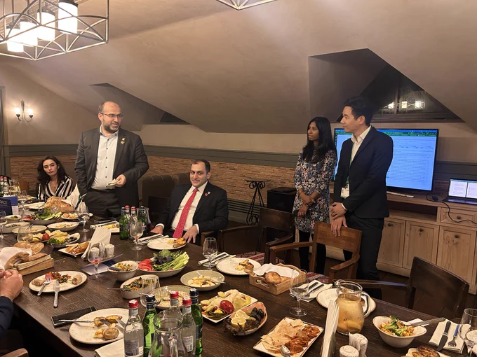 Erik Grigoryan, former Minister of Environment of Armenia, stands to speak at the dinner table. Deputy Minister Gevorg Mantashyan sits beside him, and Veena Balakrishnan and David Dao listen by a screen.
