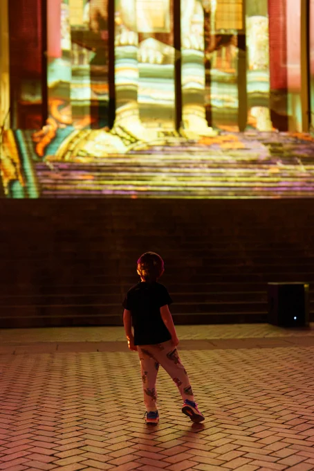 A person stands in front of the Rotunda at the University of Virginia. The Rotunda displays abstract light projections from The Great Rotumpkin.