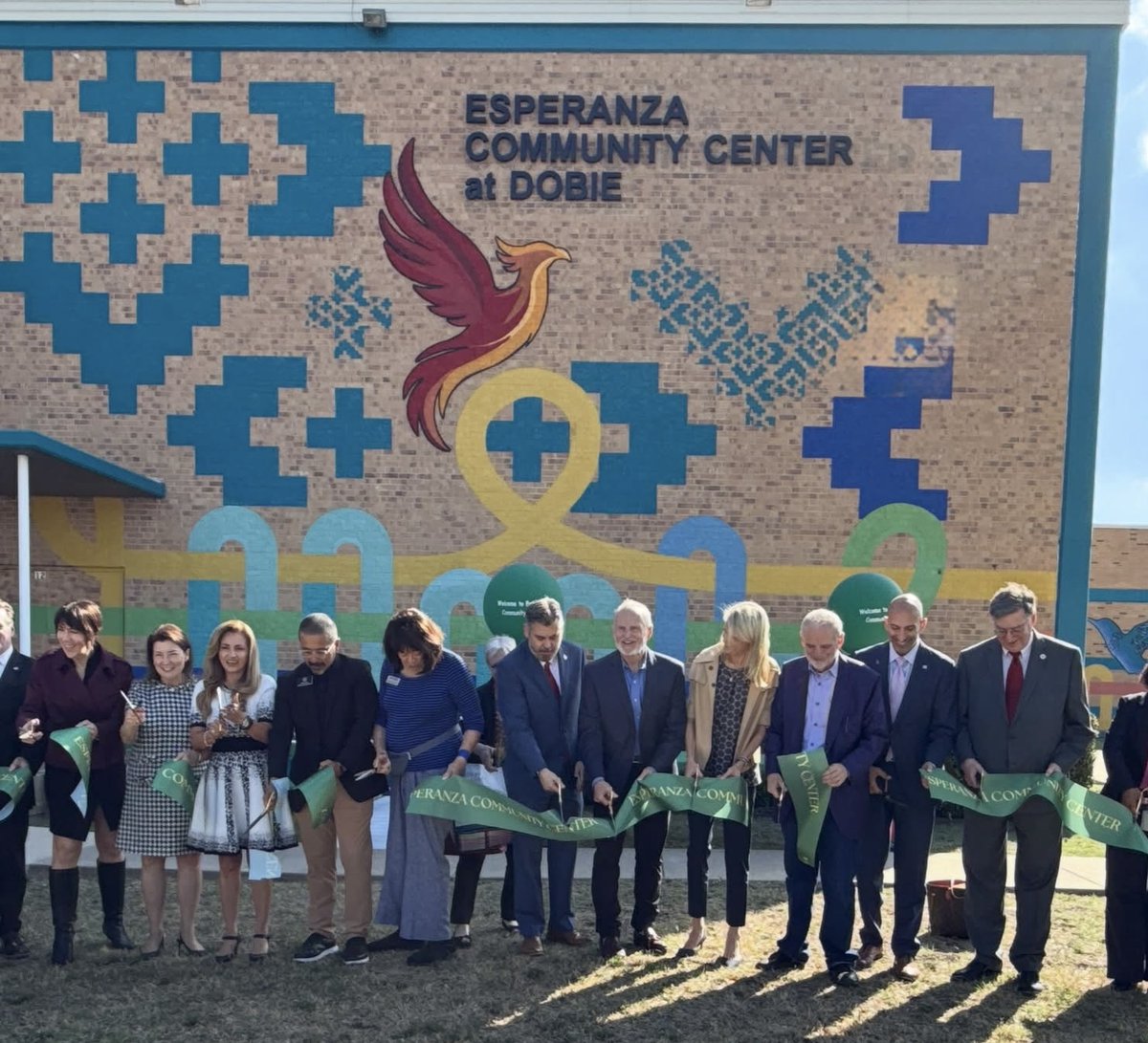 A group of people stand outside a brick community center during a ribbon-cutting ceremony, holding a green ribbon in front of a colorful mural. Text appears on the building and ribbon.