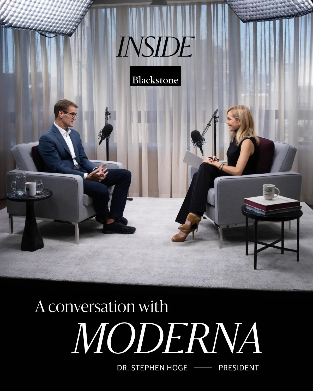 The image shows a business setting where two individuals are seated facing each other in a studio, engaged in an interview-style conversation. Both are in suits, and they are surrounded by professional recording equipment. There are coffee tables with mugs and a book next to them. The backdrop includes a curtain and two tall windows bringing in natural light. The text reads: "Inside Blackstone - A conversation with Moderna - Dr. Stephen Hoge - President.