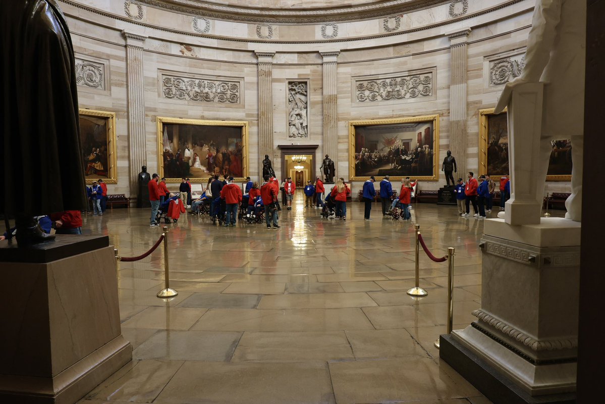 A grand, circular museum or memorial hall with marble walls, large framed paintings, statues, and a group of visitors gathered in the center. The visitors mostly wear red and blue clothing, and several are using wheelchairs. There is some text visible in the scene.