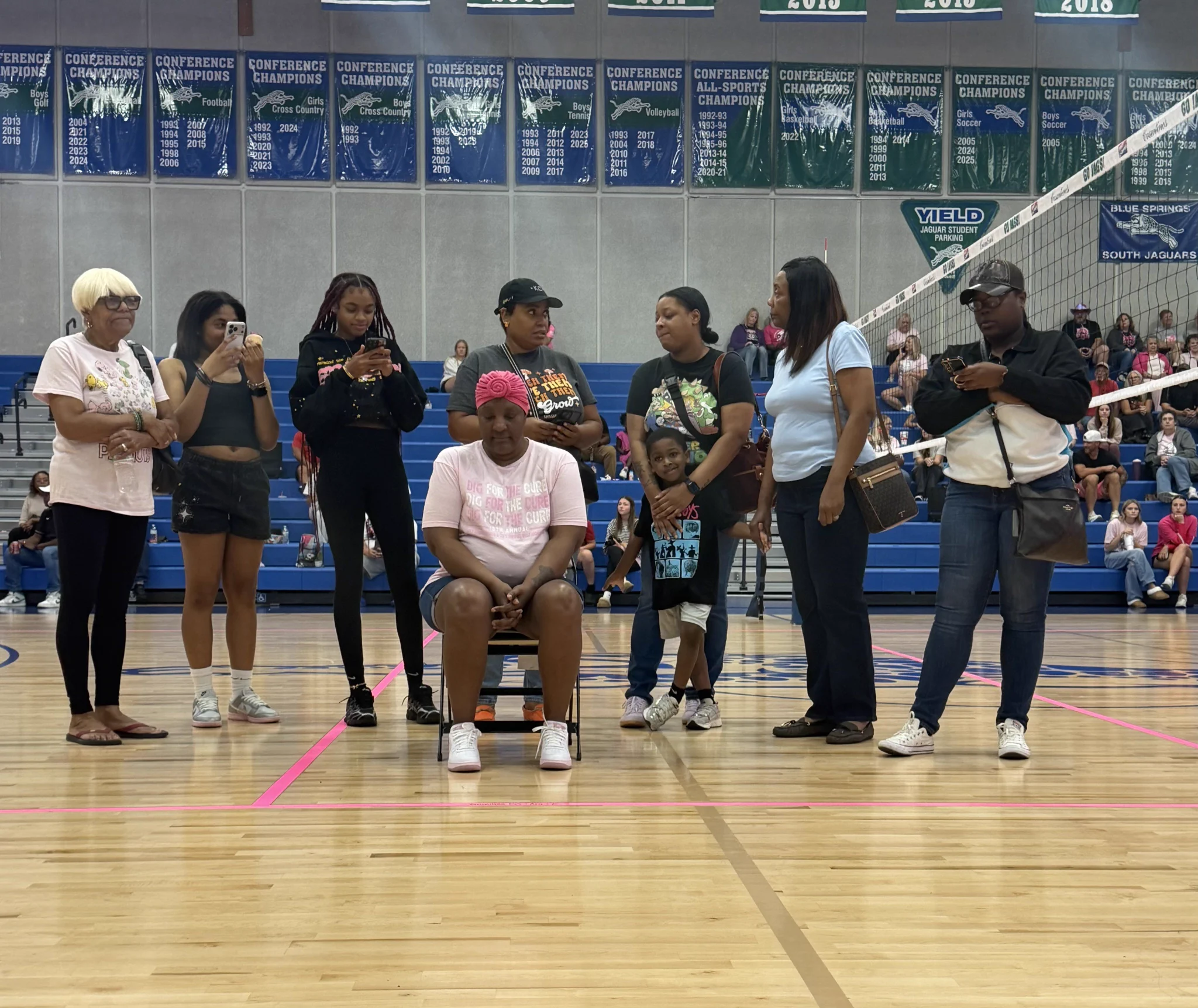 A group gathers around a seated woman in a pink shirt at a school gym.