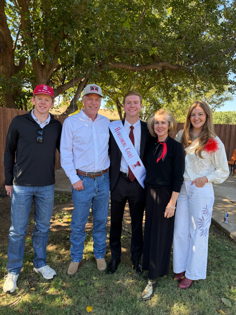 A group of five people pose together outdoors beneath a large leafy tree. The young man in the center wears a suit and a white pageant-style sash; the others stand beside him smiling. There is text on the sash and clothing.