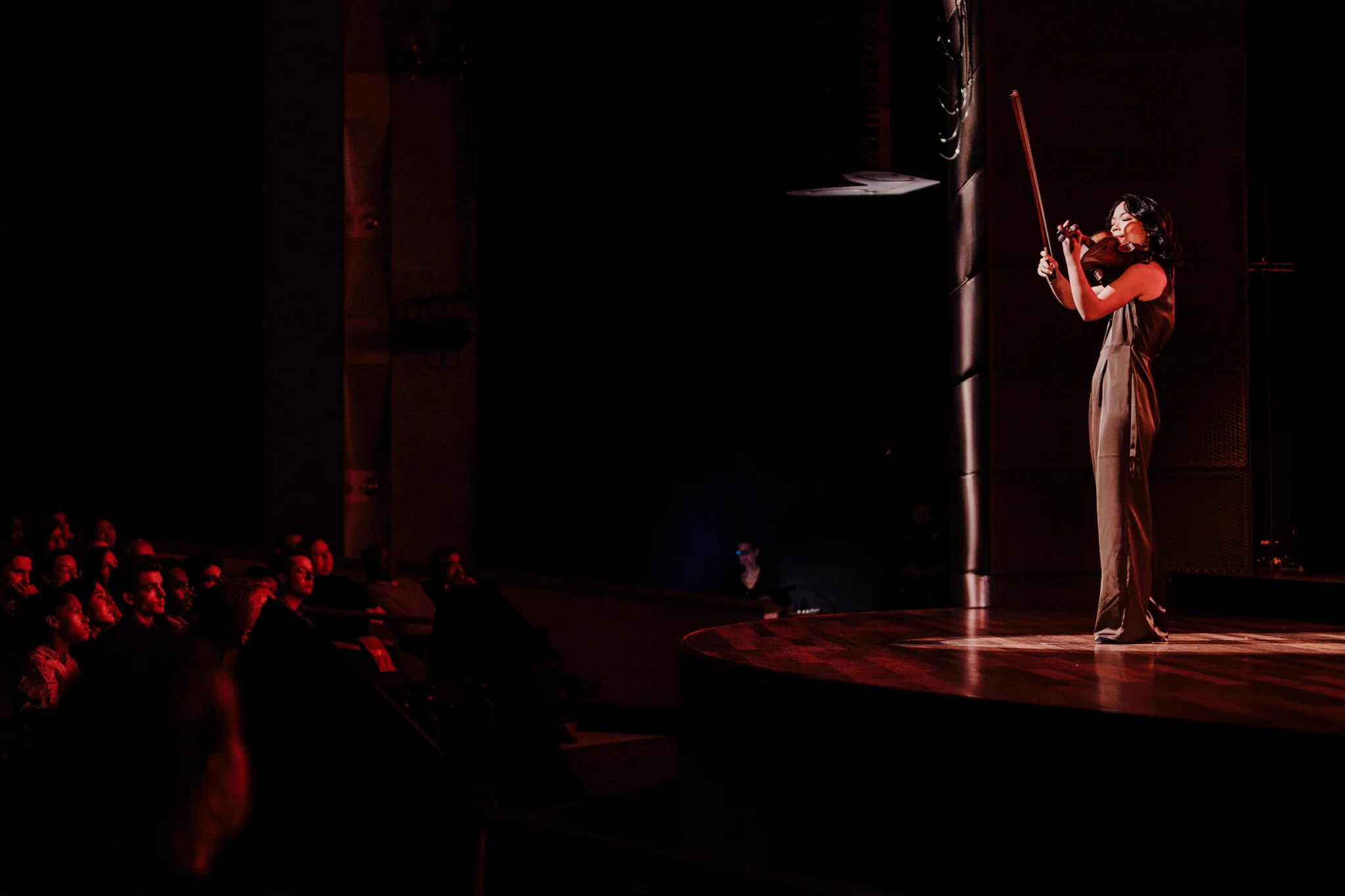 A performer in a tan jumpsuit stands on a dark stage, smiling as she plays the violin.
