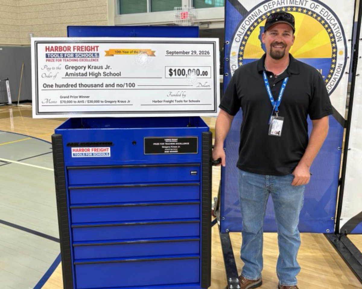 A smiling man stands beside a large blue tool chest and an oversized award check in a school gym. A California Department of Education banner is behind him. The check and banner contain text.
