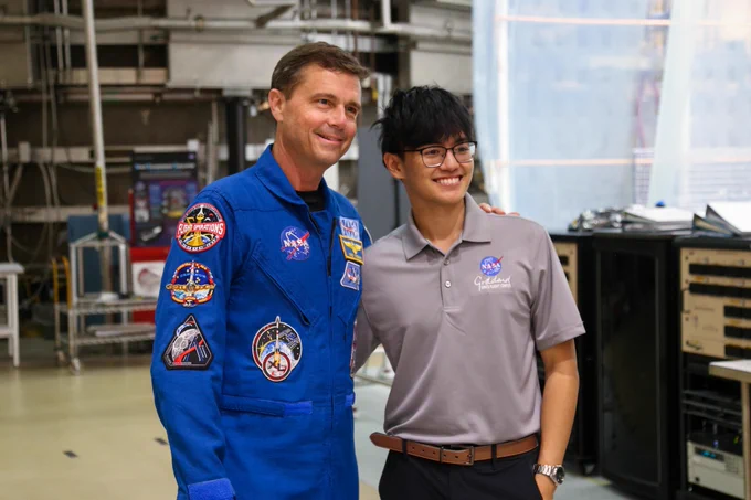 James poses for a photo alongside Reid Wiseman, who is wearing a blue flight suit, inside Goddard's Integration and Testing Facility. Both are smiling at the camera, with the Space Environment Simulator and a workstation in the background.