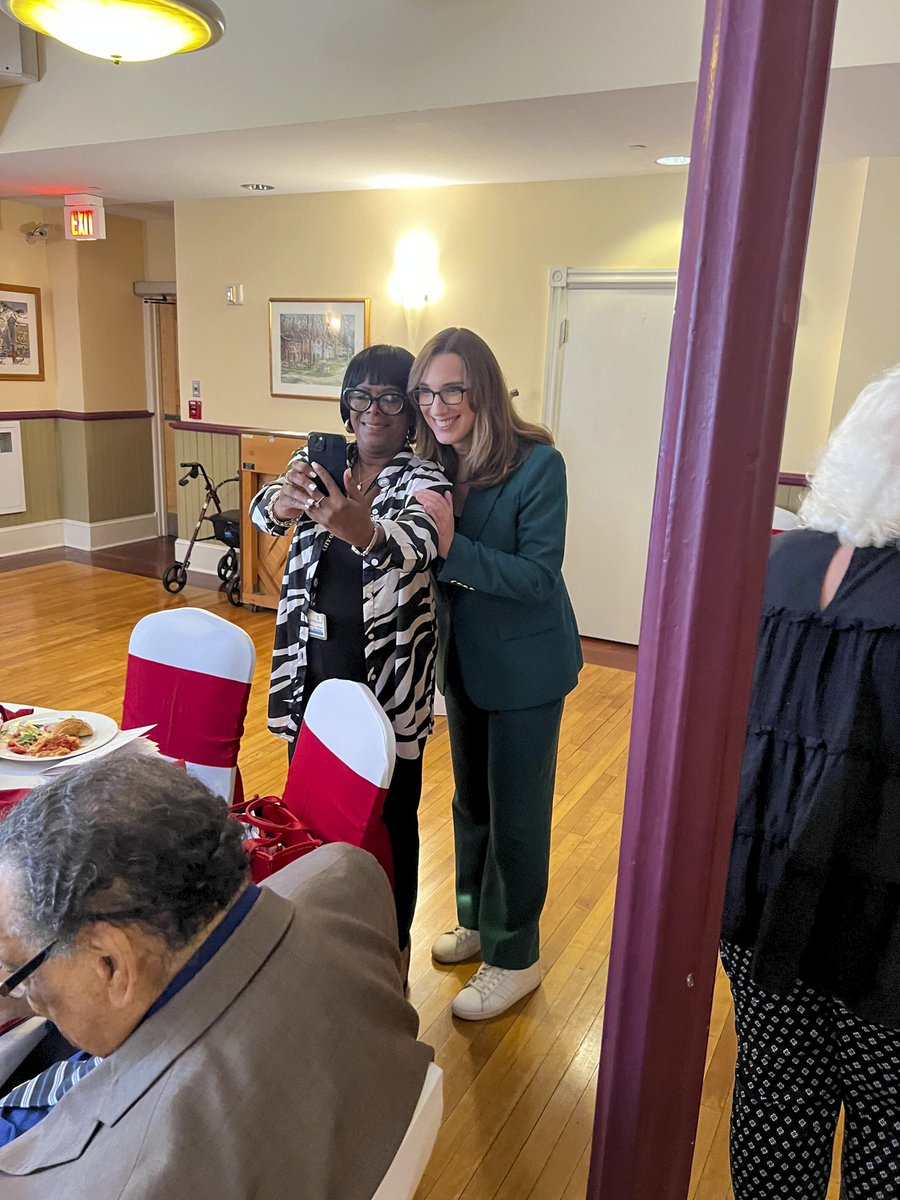 Two women stand together smiling as one takes a photo with a smartphone in a warmly lit dining or event room. Other people are seated nearby, with tables, chairs, and a walker visible. There is text on a wall sign.