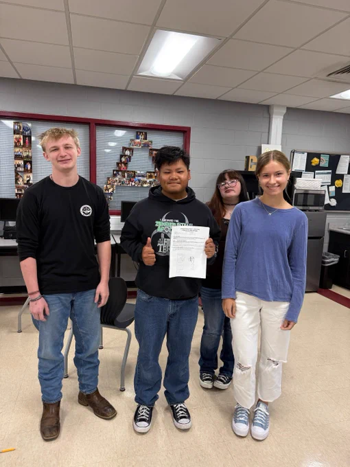Three students stand together holding their product pitch paperwork in a classroom.