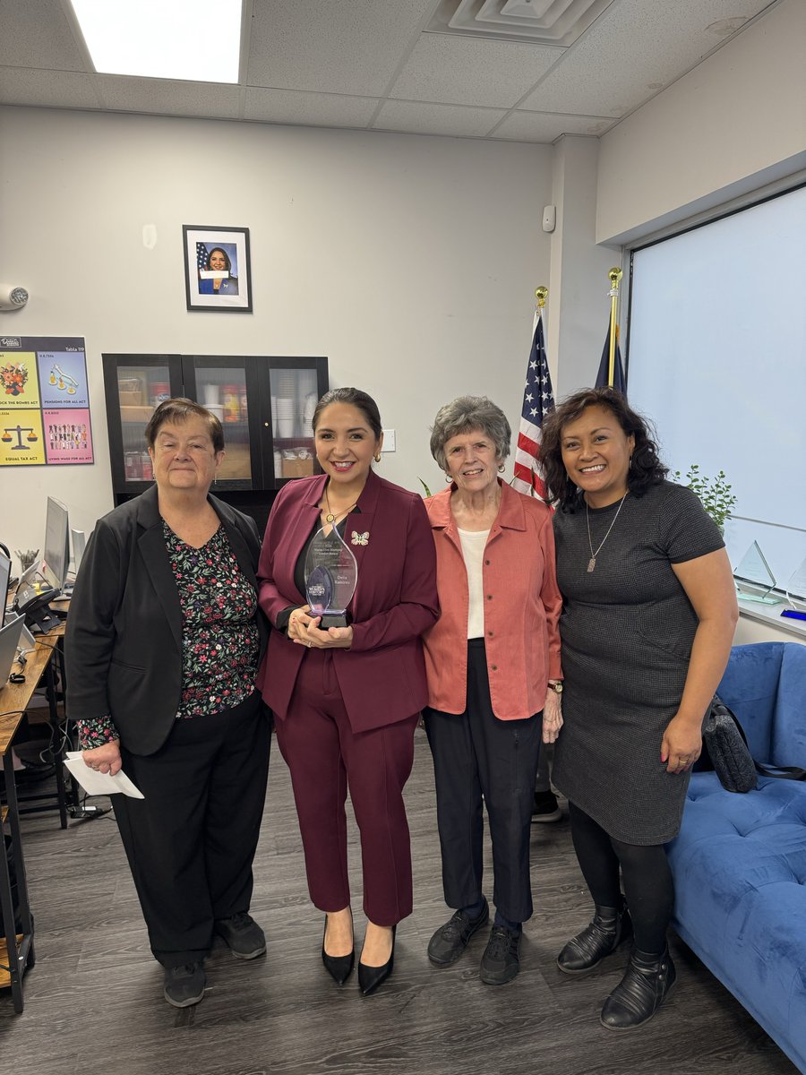 Four women pose together in an office. The woman second from the left holds a glass award. U.S. and state flags, office furniture, a blue sofa, framed picture, and wall posters are visible. There is text on the award and posters.