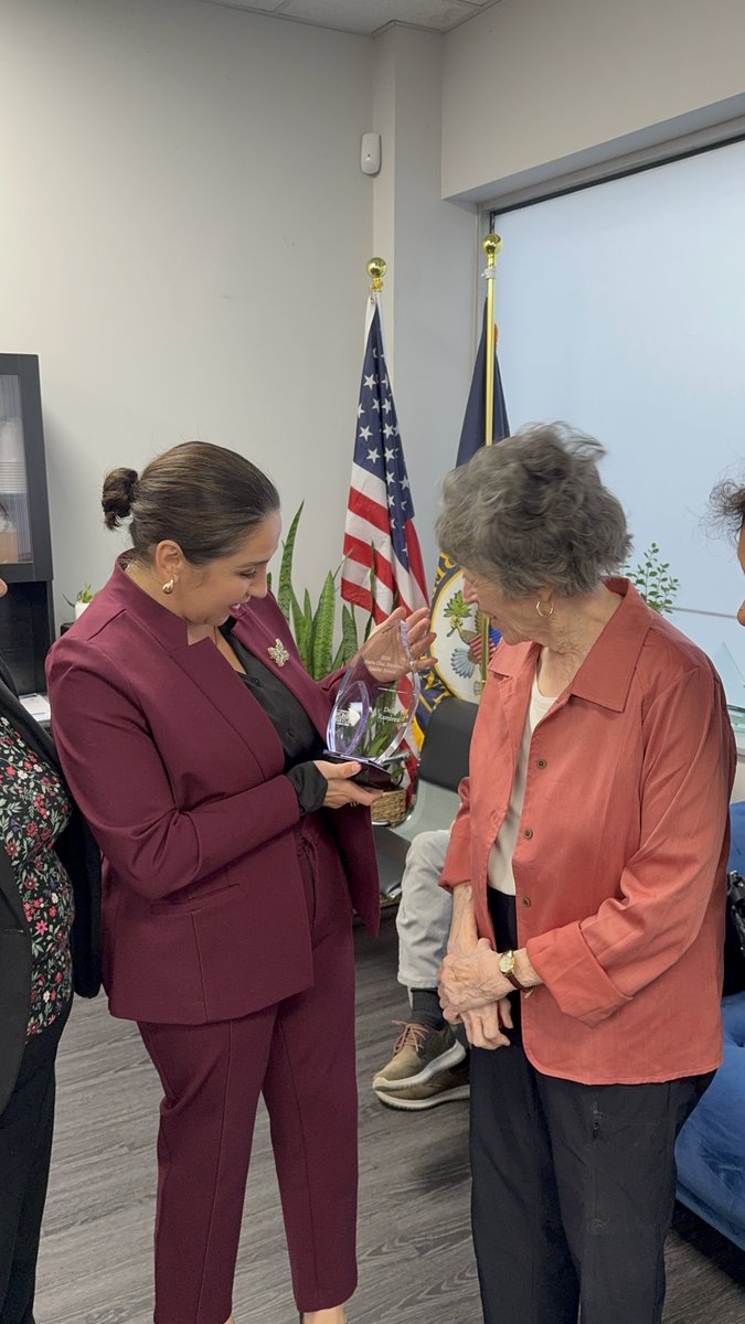 Two women stand in an office as the woman in a burgundy suit presents a clear award to an older woman in a coral shirt. Flags and several people are visible in the background. No prominent text is visible.