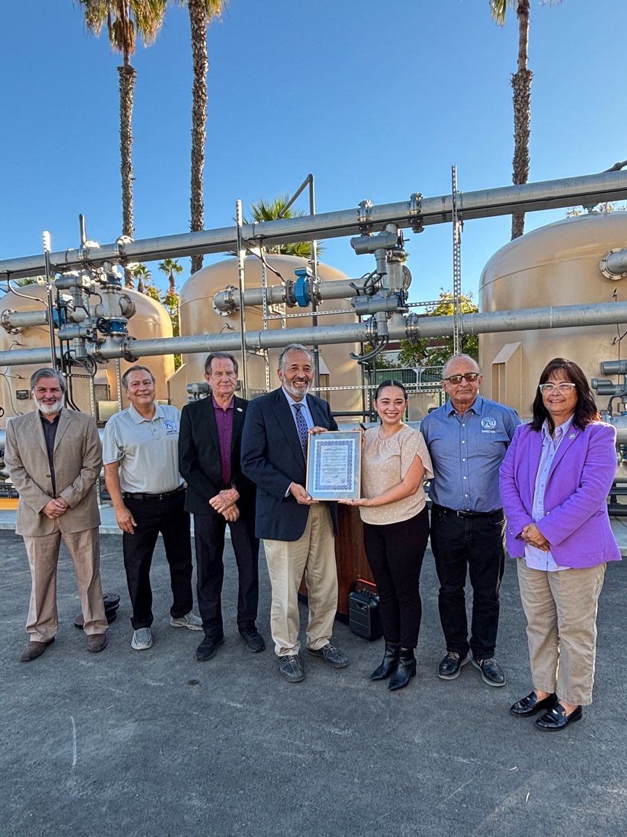 A group of seven adults pose outdoors in front of industrial tanks and pipes. Two people hold a framed certificate. Palm trees and a clear blue sky are visible in the background. The certificate contains text.