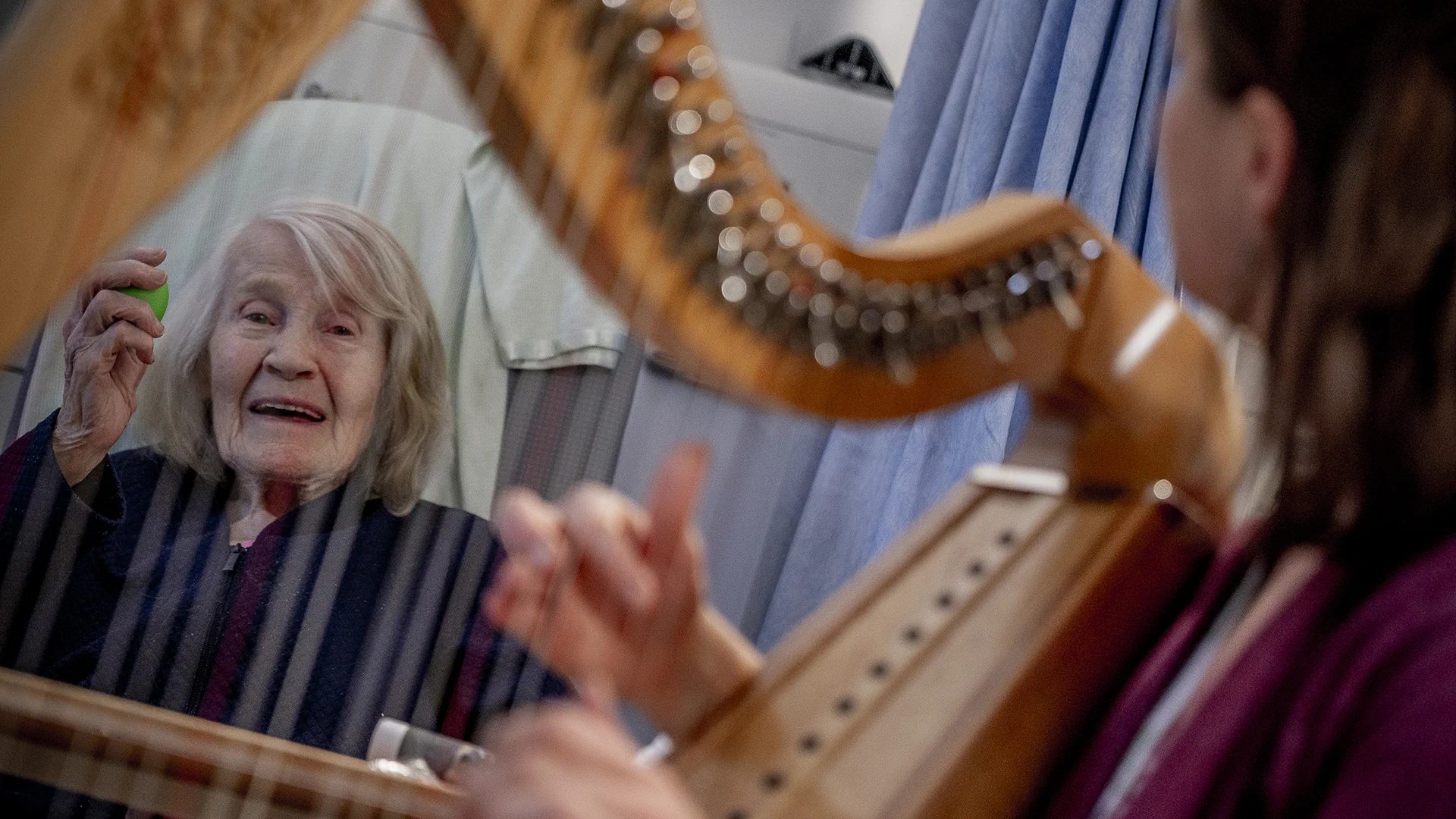 Elderly woman smiling and holding a green object while listening to a harp player in a hospital room.