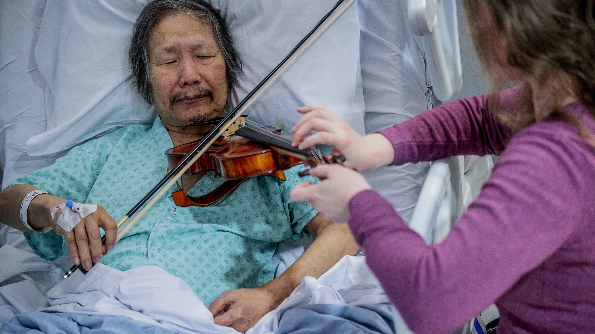 Elderly man in hospital bed playing violin with assistance from a woman in a purple sweater.
