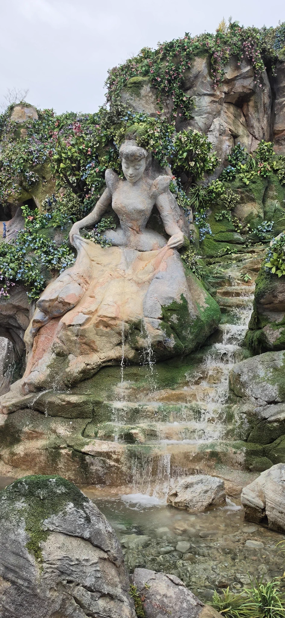 A stone sculpture of Cinderella surrounded by flowers and greenery, with water cascading down the rocks into a pool at Tokyo DisneySea.