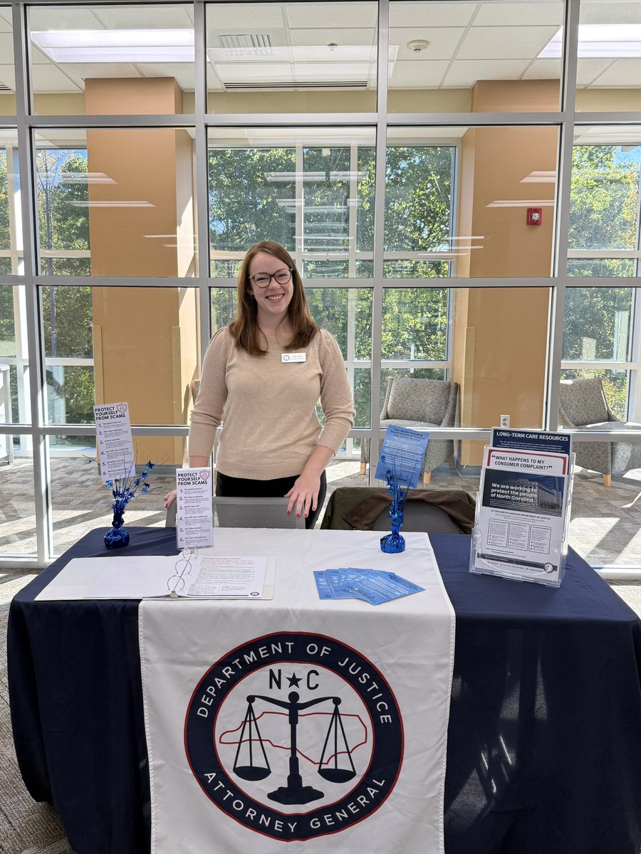 A woman stands behind an informational booth in a bright, glass-walled indoor space. The table displays pamphlets, signs, and a large government seal. There is text on the signs, pamphlets, and tablecloth.