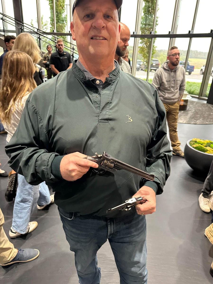 A man wearing a cap, dark green pullover, and jeans stands indoors among other people, holding two antique-looking handguns. There is a logo on his pullover, but no prominent text.
