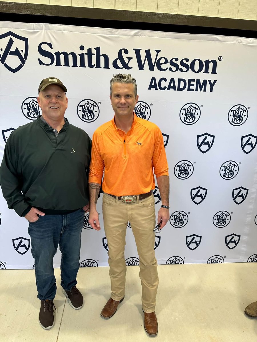 Two men pose for a photo in front of a branded event backdrop. The backdrop displays large text and repeating logos.