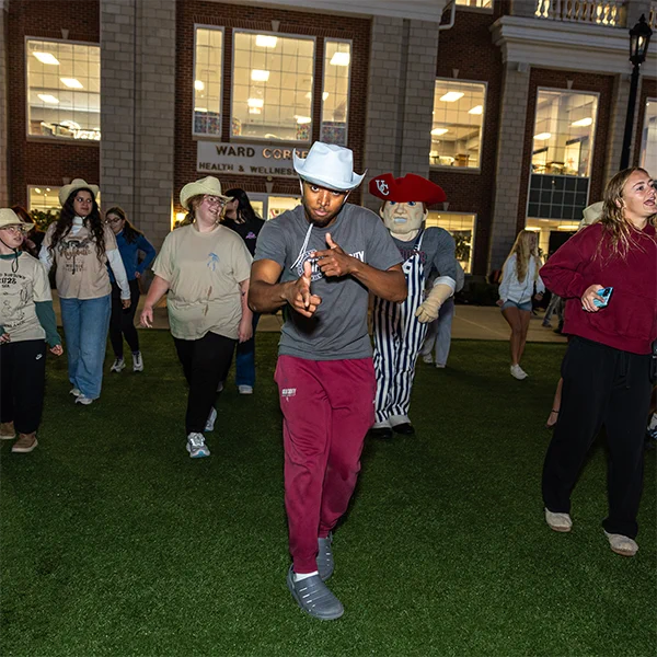 University of the Cumberlands student wearing a grey shirt, red pants, and a white cowboy hat points at the camera during Hoco Hoedown on the wellness lawn.